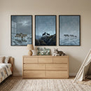 Three large framed prints of wolves in snowy landscapes hang above a light-colored wooden dresser in a serene bedroom. The artworks show wolves on icy terrain and rocky peaks under moody, clouded skies. A small, textured lamp, two nature-themed books, and a plush wolf toy sit atop the dresser. To the sides, a corner of a bed with neutral bedding and a cream canvas teepee complete the cozy, wilderness-inspired decor. A textured light-colored rug covers the floor.