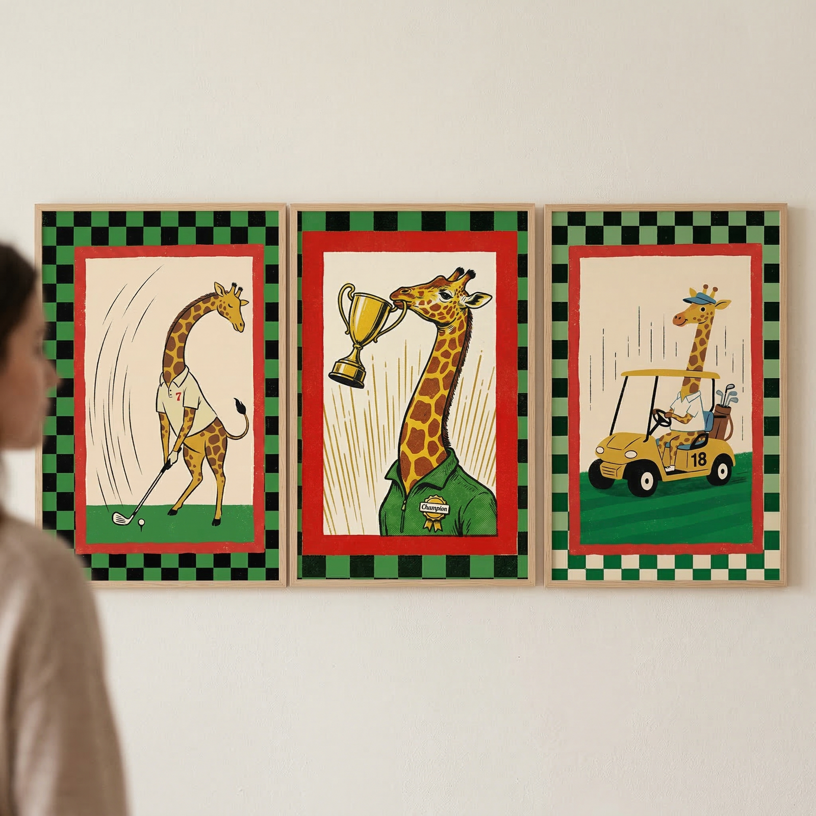 Three framed illustrations are mounted side-by-side on a plain wall, with a person's shoulder and head visible on the left side of the frame. Each of the three pieces features a cartoon giraffe and has a matching green and black checkered border surrounding a red inner frame.
The illustration on the left shows a giraffe in a white shirt bent over, preparing to hit a golf ball with a club. Curved lines suggest the motion of a swing. 