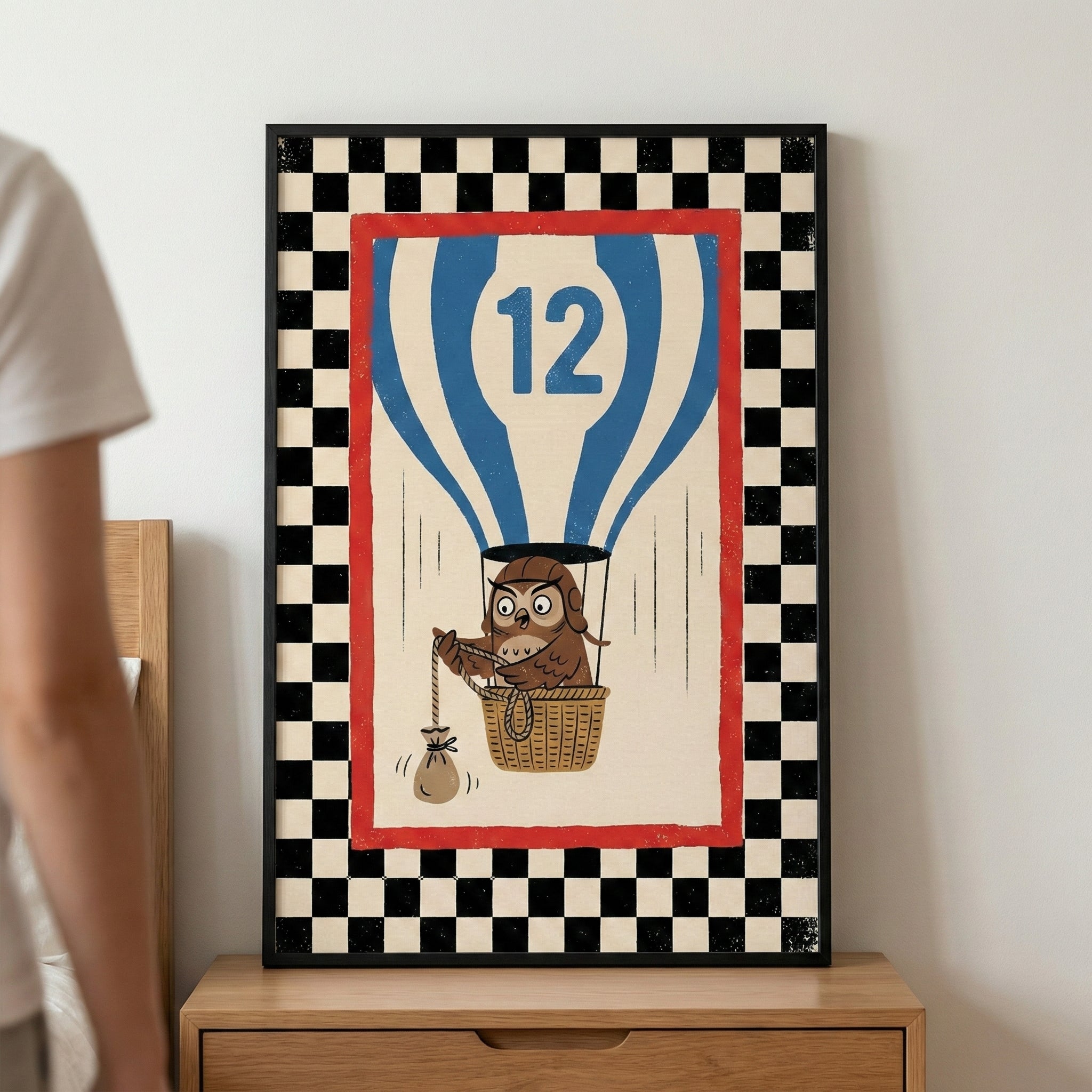 A framed graphic illustration resting on a wooden bedside chest. The artwork features a brown cartoon owl wearing aviator goggles, sitting in a woven basket attached to a blue and white striped hot air balloon. The balloon prominently displays the number "12." The owl holds a rope tied to a small sandbag hanging outside the basket. The central image is surrounded by a thin red border, all set within a wide border of black and white checkers.