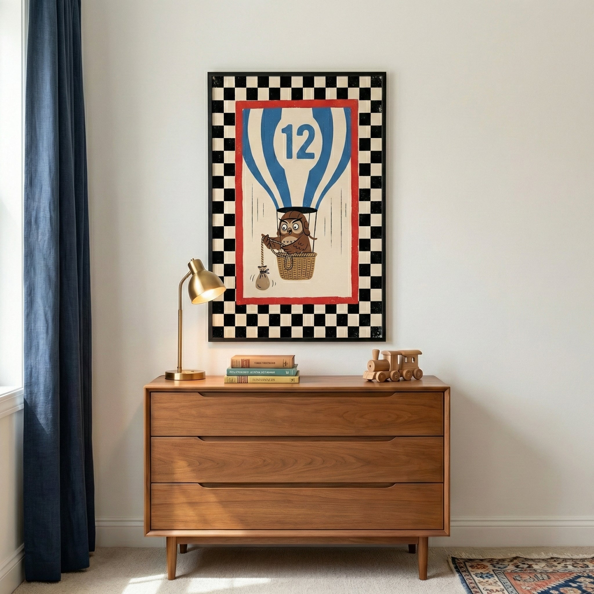 A framed art print hangs on a white wall above a three-drawer wooden dresser. The artwork depicts a brown cartoon owl wearing aviator goggles sitting in the basket of a blue and white striped hot air balloon marked with a large blue number 12. The central image is enclosed within a red rectangular border and an outer black and white checkered border. On the dresser sits a brass desk lamp, a small stack of books, and a wooden toy train. 