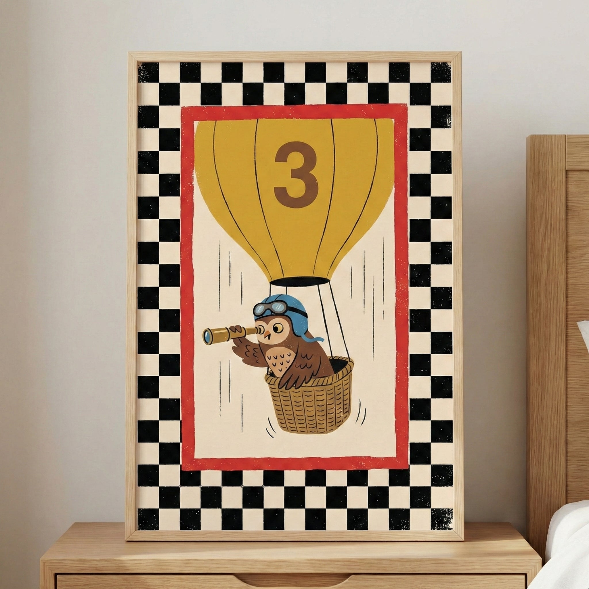 A framed illustration of a cartoon owl in a hot air balloon. The owl wears a blue aviator helmet with goggles and looks through a gold telescope. The yellow hot air balloon features a large brown number three. The central image is set against an off-white background with a red border, surrounded by a wide black and white checkerboard pattern. The artwork is housed in a light wood frame and sits on a wooden bedside table.