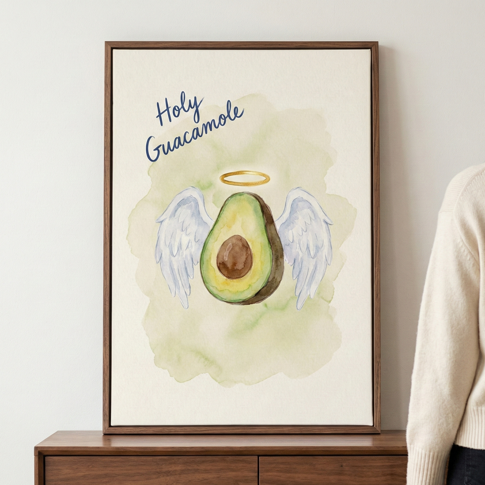 A framed vertical wall hanging featuring a watercolor-style illustration of an avocado half with its brown pit, white feathered wings, and a golden halo. The phrase "Holy Guacamole" is written in blue script above the avocado. The illustration is set against a pale green wash on a cream-colored background. The thin, dark wood frame is mounted on a white wall above a brown wooden dresser. A person wearing a cream-colored sweater is partially visible on the right side of the image.