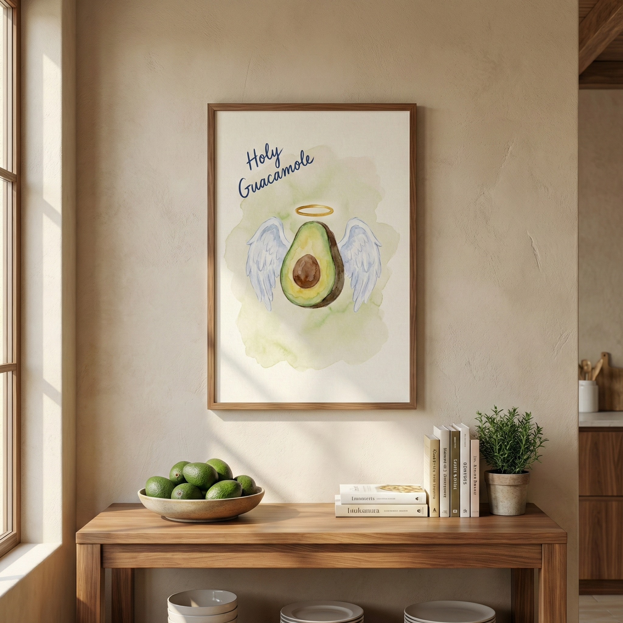 "A framed art print hangs on a textured, light beige wall above a wooden console table. The print features a whimsical watercolor illustration of a halved avocado with a brown pit, sprouting white feathered wings and topped with a thin golden halo. Above the avocado, the words ""Holy Guacamole"" are written in a playful, dark blue cursive script. The central figure is surrounded by a soft, light green watercolor wash. 
