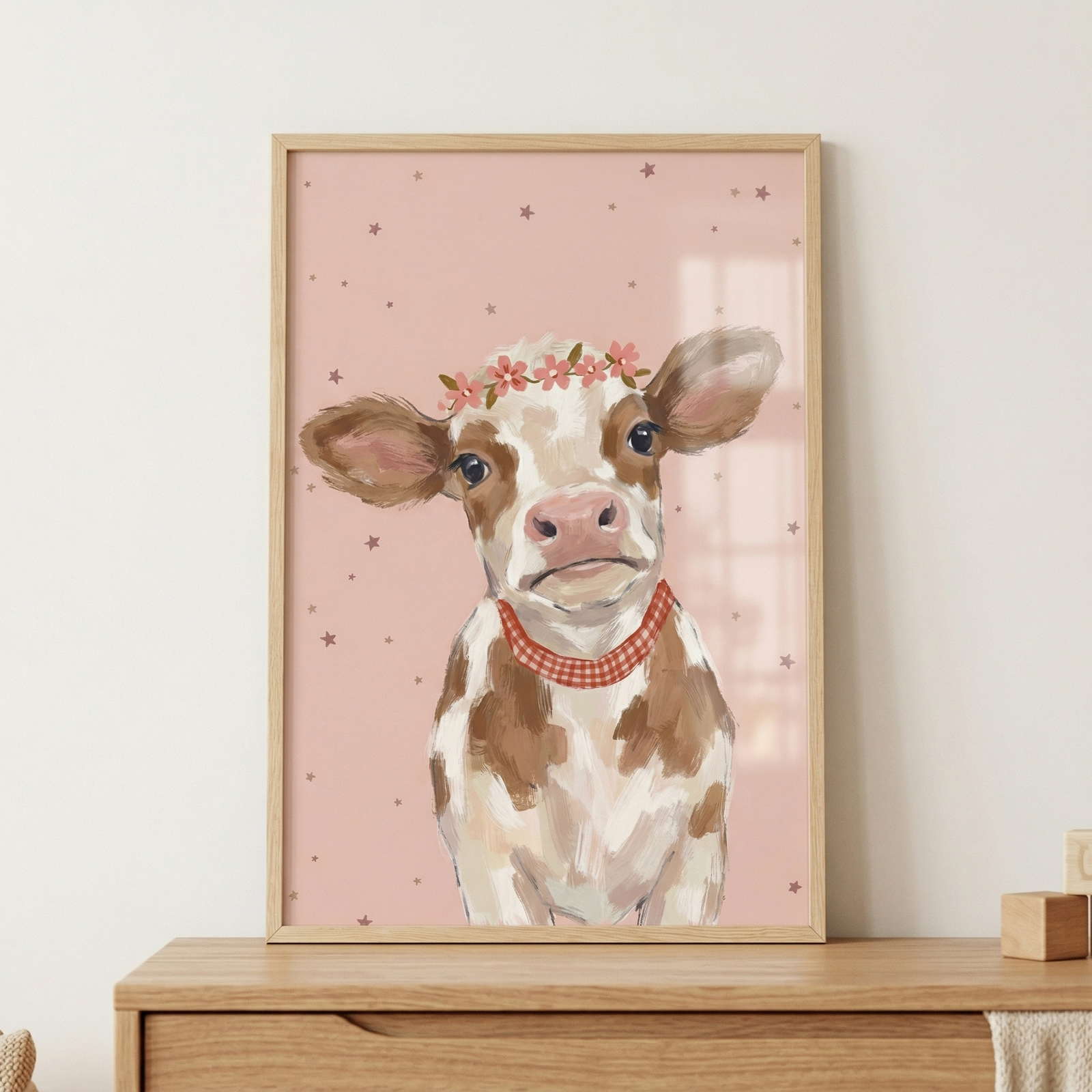 A framed illustration of a brown and white calf sits on a wooden dresser. The calf wears a crown of small pink flowers and a red-and-white checkered bandana. It is set against a soft pink background scattered with tiny brown stars. The artwork is enclosed in a light-toned wood frame. The overall style is whimsical and gentle. This charming depiction of a young animal is presented in a minimal, modern setting.