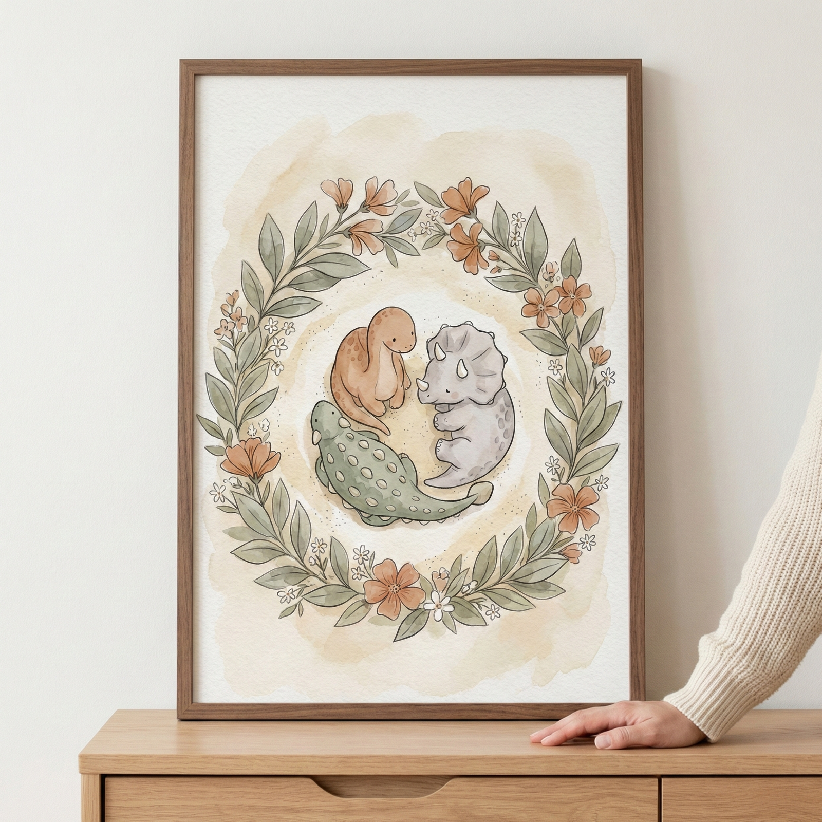 A vertical wood-framed watercolor illustration featuring three cute, stylized dinosaurs nestled within a circular wreath of muted green leaves and soft orange flowers. The group includes a peach-colored long-necked dinosaur, a light gray horned dinosaur, and a green armored dinosaur. The artwork is set against a warm, wash-like background and is resting on a light-toned wooden dresser. A person's hand is visible touching the bottom right corner of the frame.