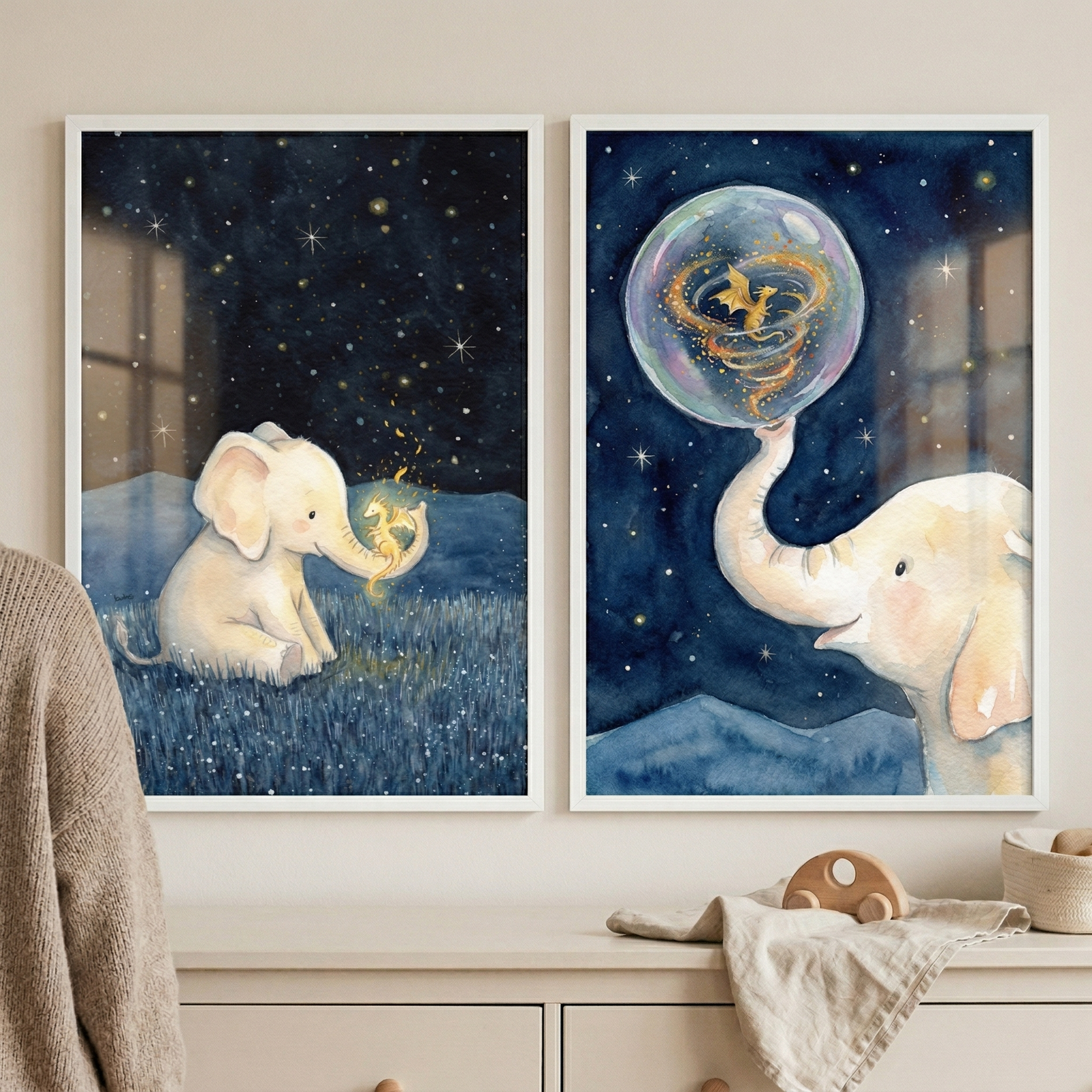 Two framed watercolor-style artworks hang side-by-side on a light-colored wall above a wooden dresser. Both pieces feature a whimsical, dreamy theme set against a dark blue night sky filled with twinkling stars.
In the left frame, a small, pale-colored elephant sits in a field of deep blue grass. It holds a tiny, glowing golden dragon in its trunk. The scene is calm and magical, with soft highlights on the elephant’s skin.

