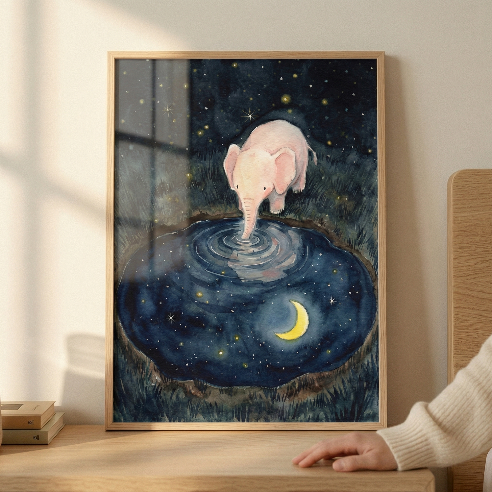 A framed illustration depicts a small, pale pink elephant standing at the edge of a circular pond at night. The elephant dips its trunk into the center of the water, creating subtle ripples. The pond acts as a mirror, reflecting a deep blue sky filled with numerous small stars and a bright yellow crescent moon. The ground surrounding the water is covered in dark, textured grass. 
