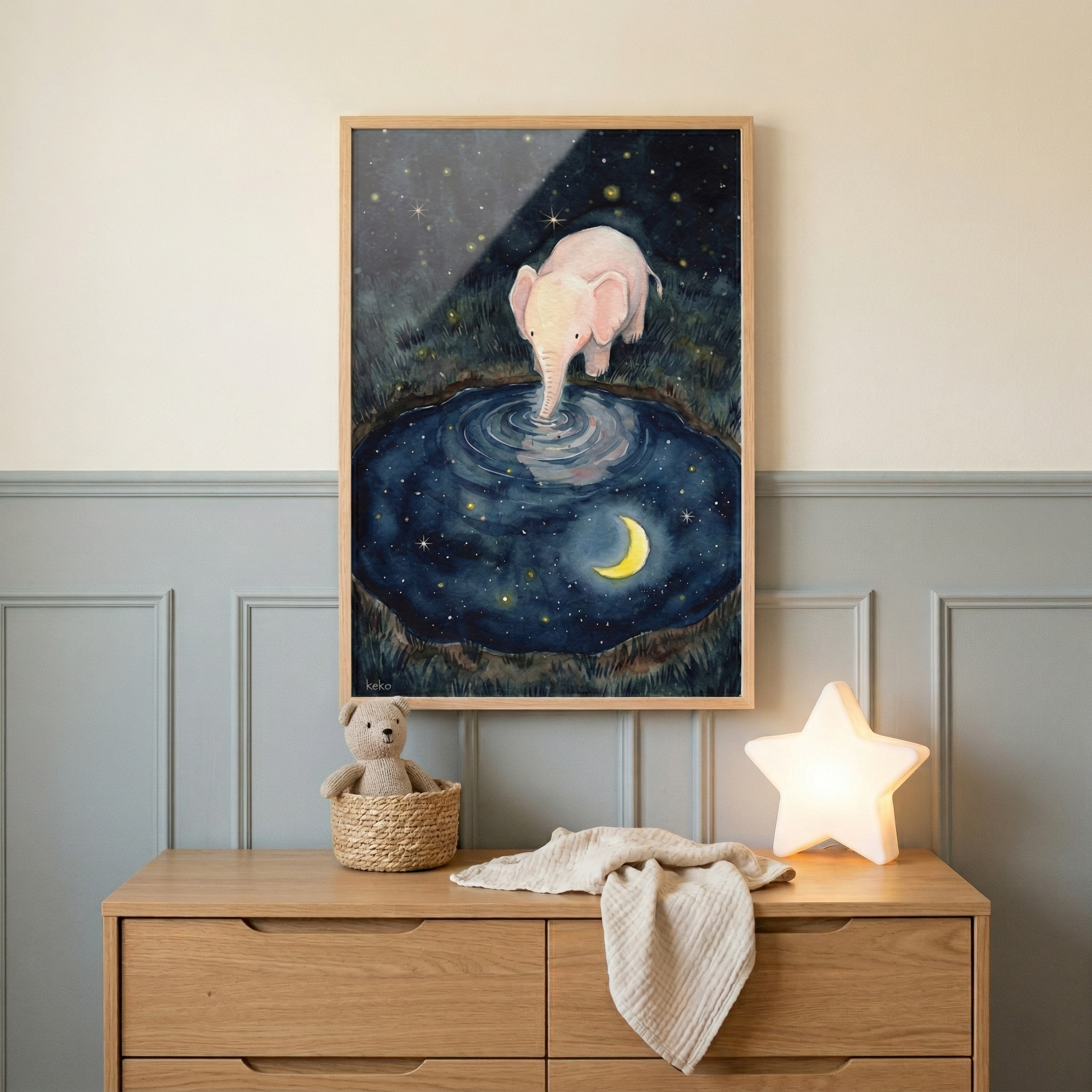 A framed rectangular artwork hangs on a wall above a light wood dresser. The artwork depicts a small, light pink cartoon elephant standing by a circular pool of water at night. The elephant's trunk is dipped into the water, which reflects a yellow crescent moon and numerous tiny stars, mirroring the starry night sky above. Below the frame, a wooden dresser sits against a wall with light blue-grey paneling. 