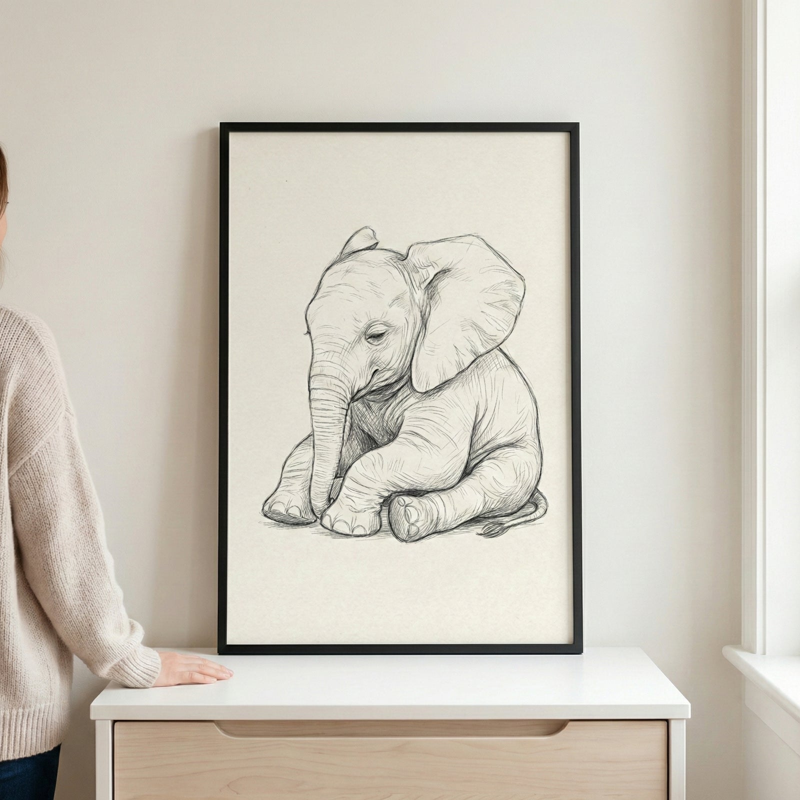 A black-framed pencil sketch of a baby elephant sitting down, resting on a light-colored wooden dresser against a neutral wall. A person's arm and hand are visible on the left, resting on the surface next to the artwork.
