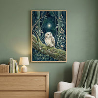 A framed vertical illustration hangs on a sage green wall in a cozy room setting. The artwork depicts a small, fluffy owl perched on a mossy tree branch in a dark, whimsical forest at night. The owl looks upward toward a single bright star shining in the deep blue sky. Surrounding the owl are delicate leaves, ferns, and small white flowers. Below the artwork sits a light wood dresser holding a small white lamp, a row of books, and a woven basket. To the right, a beige armchair is draped with a textured gree