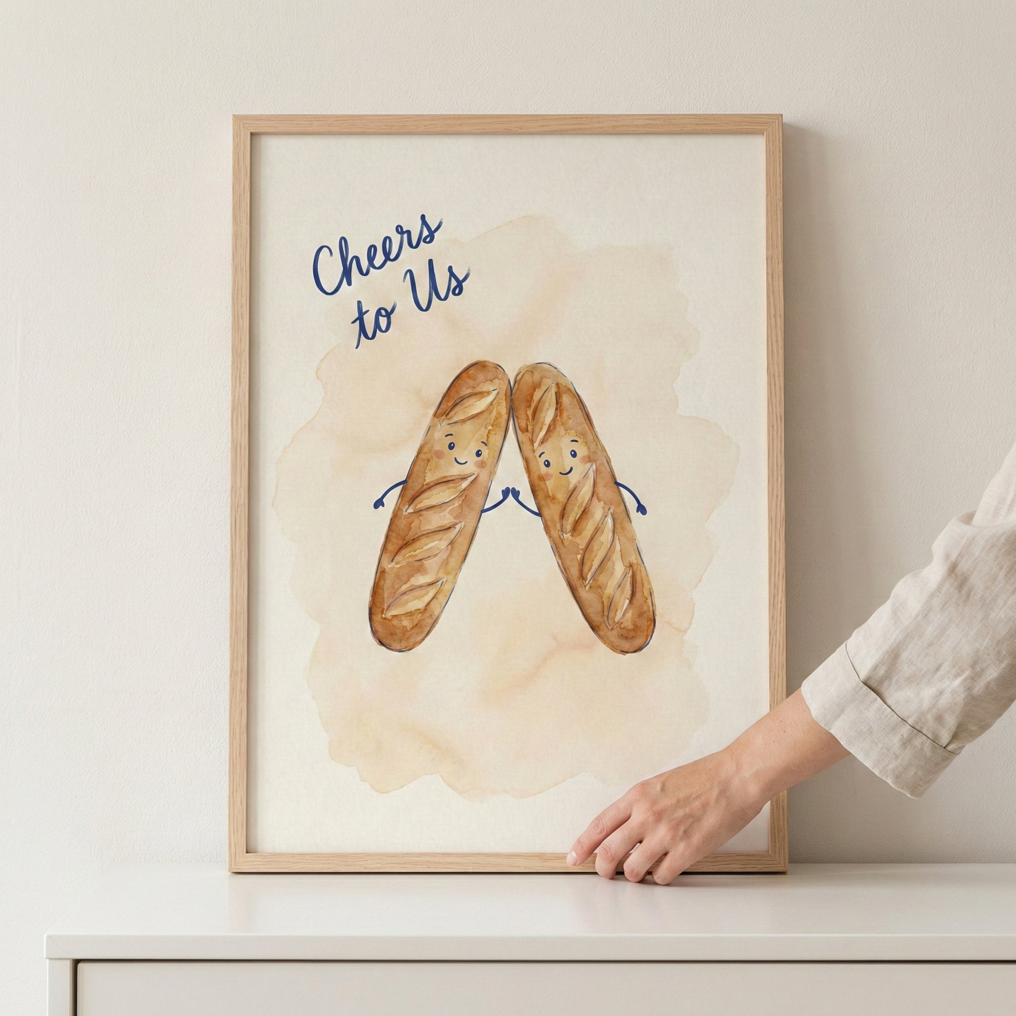A framed illustration shows two smiling cartoon baguettes with tiny blue arms clinking together as if making a toast. The text "Cheers to Us" is written above them in a dark blue script. The artwork is set against a soft, light-tan watercolor-style wash. A hand is touching the bottom edge of the light wood frame, which sits on a white surface against a plain wall.