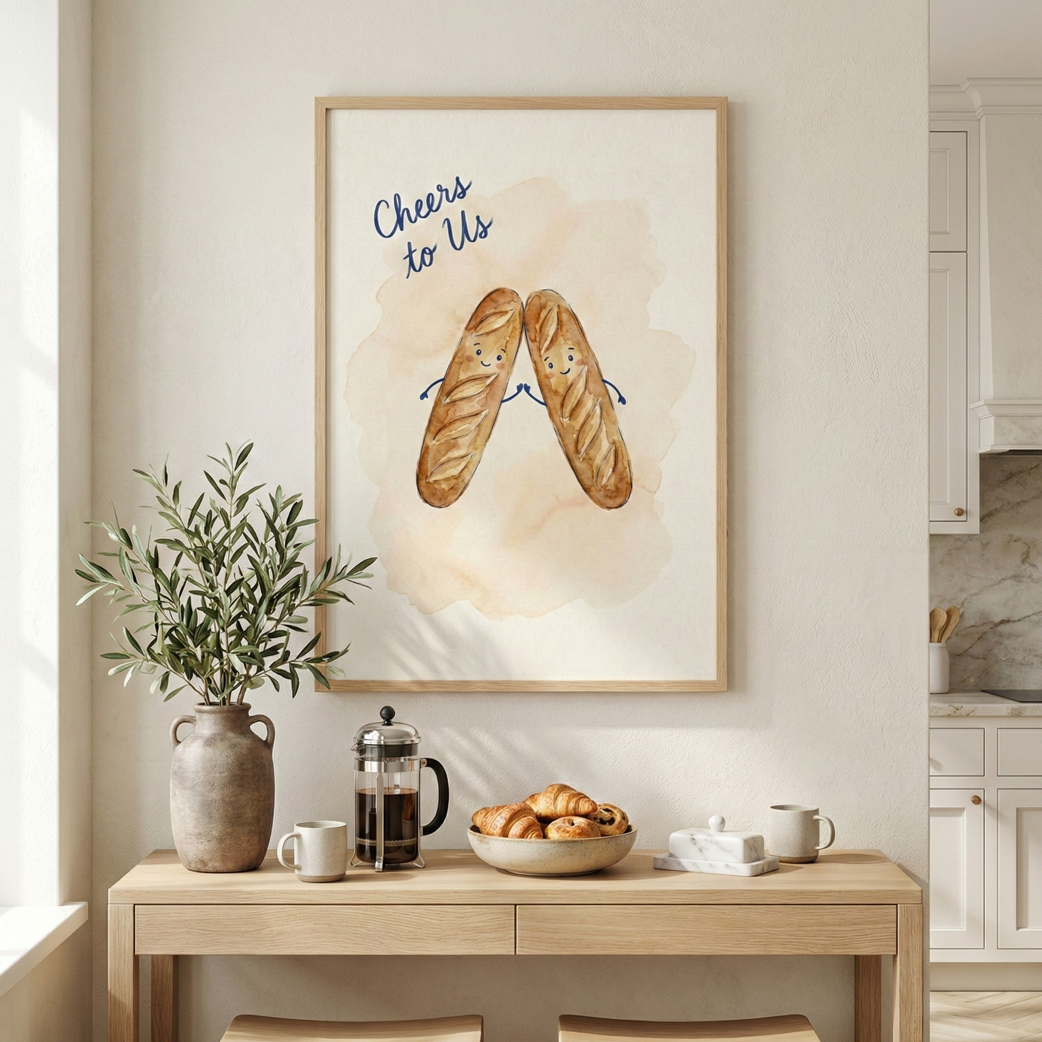 A framed piece of wall art hangs on a light-colored wall above a wooden console table. The illustration inside the frame features two cheerful, cartoon baguettes with smiling faces clinking together in a celebratory gesture. Above the characters, the phrase "Cheers to Us" is written in a navy blue script. The illustration is set against a soft, warm-toned watercolor wash. 