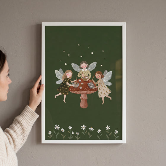 A woman holds a framed baking mushroom garden fairies wall art print showing fairies baking on a large mushroom cap.