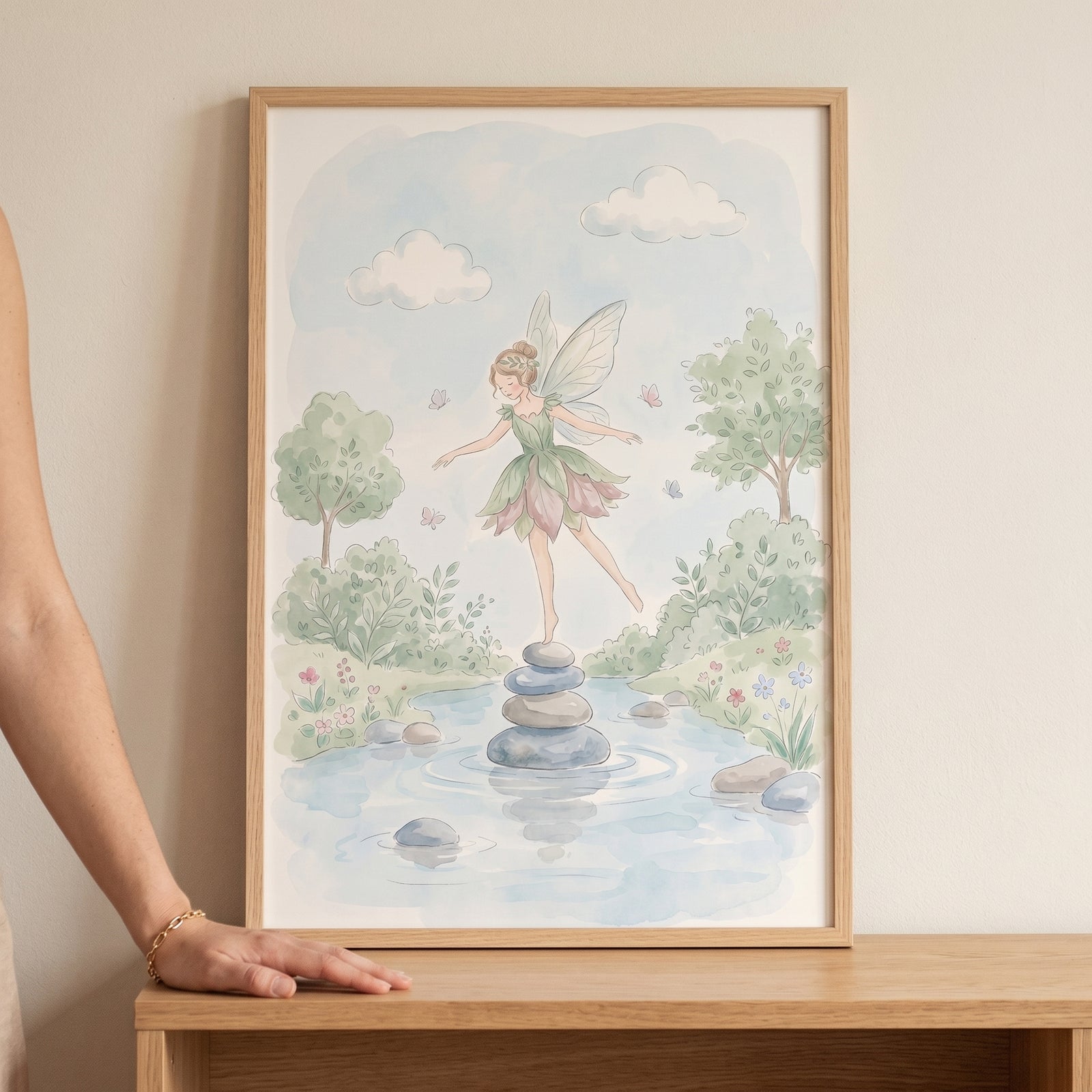 Framed balancing stone fairy wall art of a winged fairy on stones in a pond with soft trees and clouds in watercolor style.