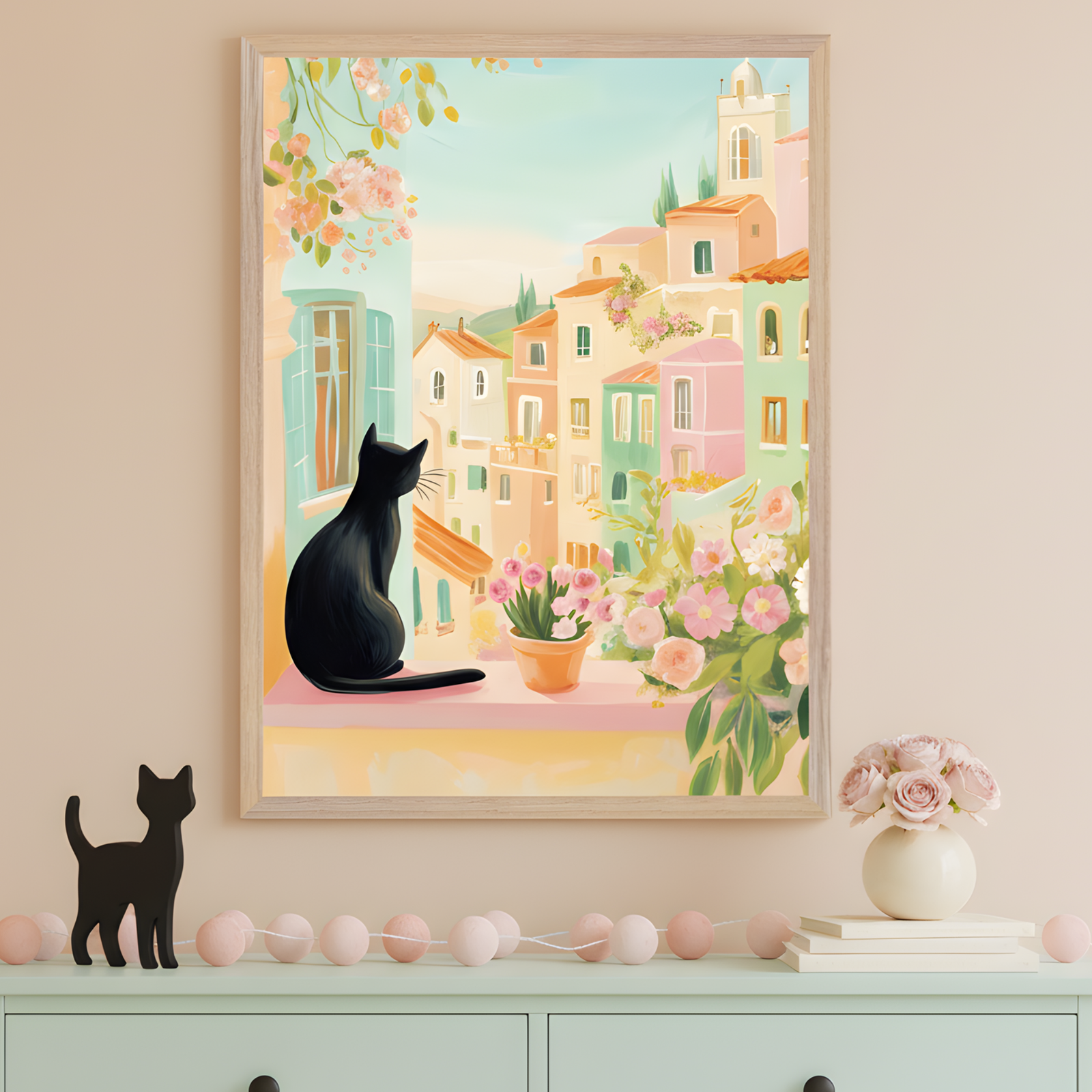 An illustration of a black cat sitting on a windowsill overlooks a pastel-colored Mediterranean village, with terracotta rooftops and blooming flowers. The scene is soft and dreamy, with pink and peach blossoms in pots and climbing vines framing the view. Below the artwork, a mint green dresser is decorated with a small black cat figurine, pink garland, and a vase of roses.