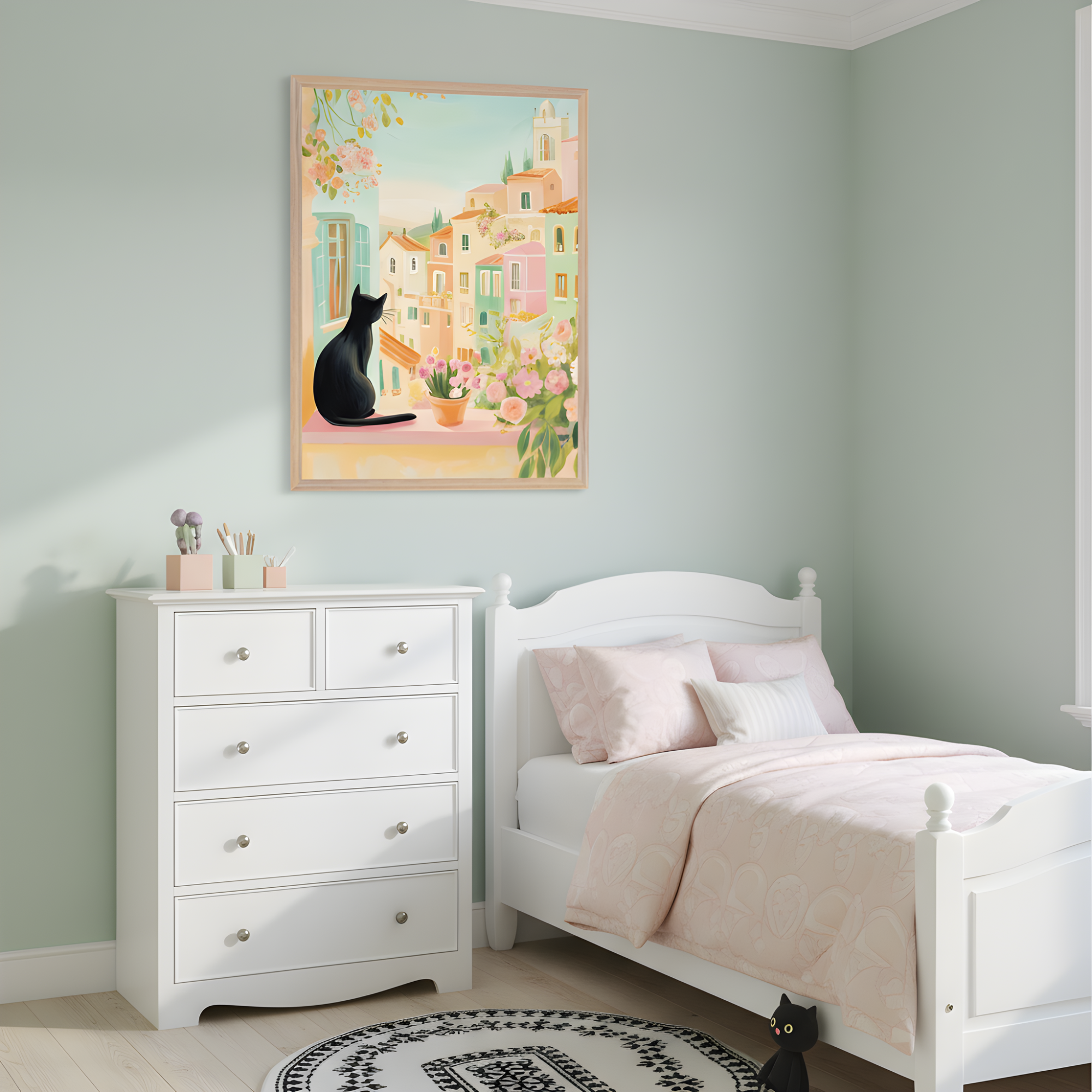 Pastel-themed child's bedroom features a white bed with soft pink bedding, a white dresser with art supplies, and a circular black-and-white rug. Above the dresser, a framed illustration shows a black cat sitting on a window ledge overlooking a sunlit Mediterranean village filled with colorful buildings and blooming flowers. A small black cat figurine on the floor subtly echoes the artwork above.