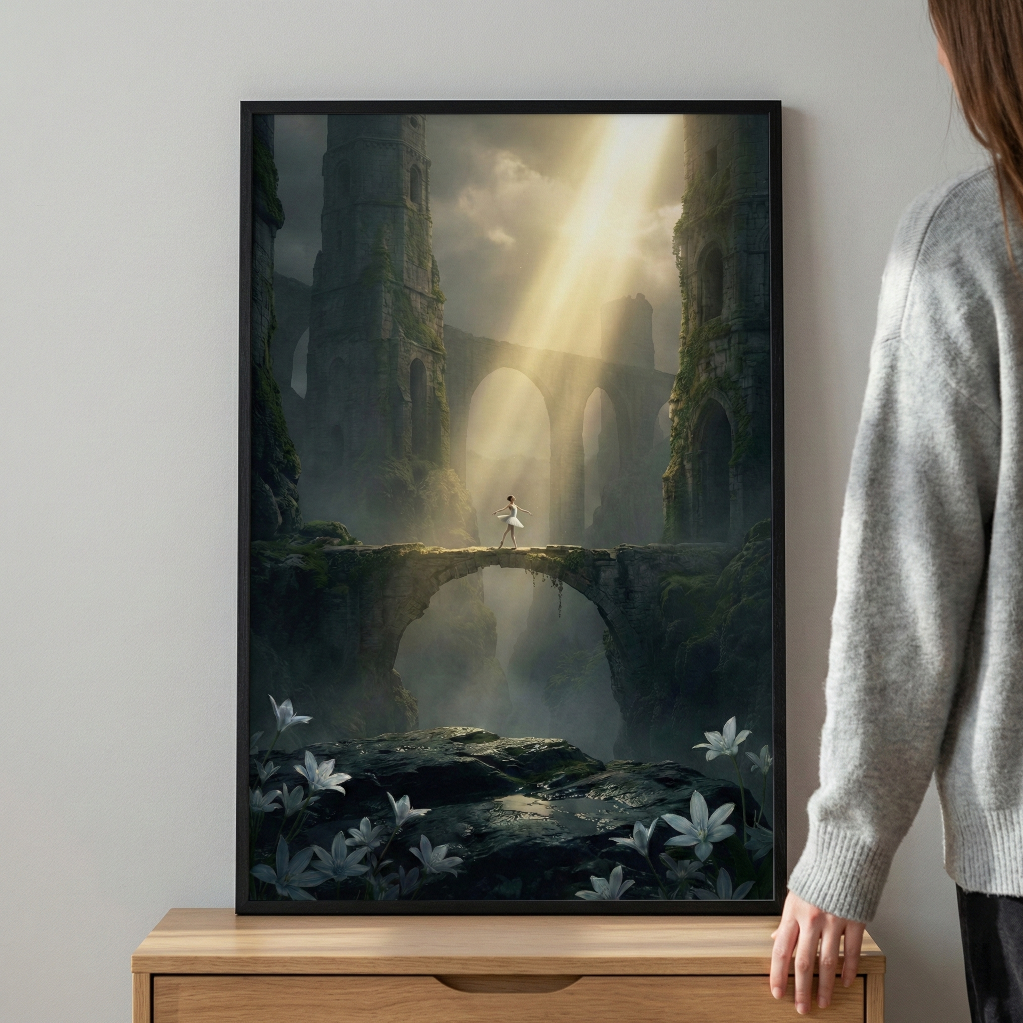 A framed art print sits on a wooden cabinet, leaning against a neutral wall with a person in a gray sweater standing beside it. The artwork depicts a small dancer in white walking across an ancient stone bridge. A bright, ethereal beam of golden light shines down from a cloudy sky, illuminating the figure. Massive, ivy-covered stone towers and arches rise from a misty valley in the background. In the foreground, large white lilies bloom among dark, shadowed rocks.