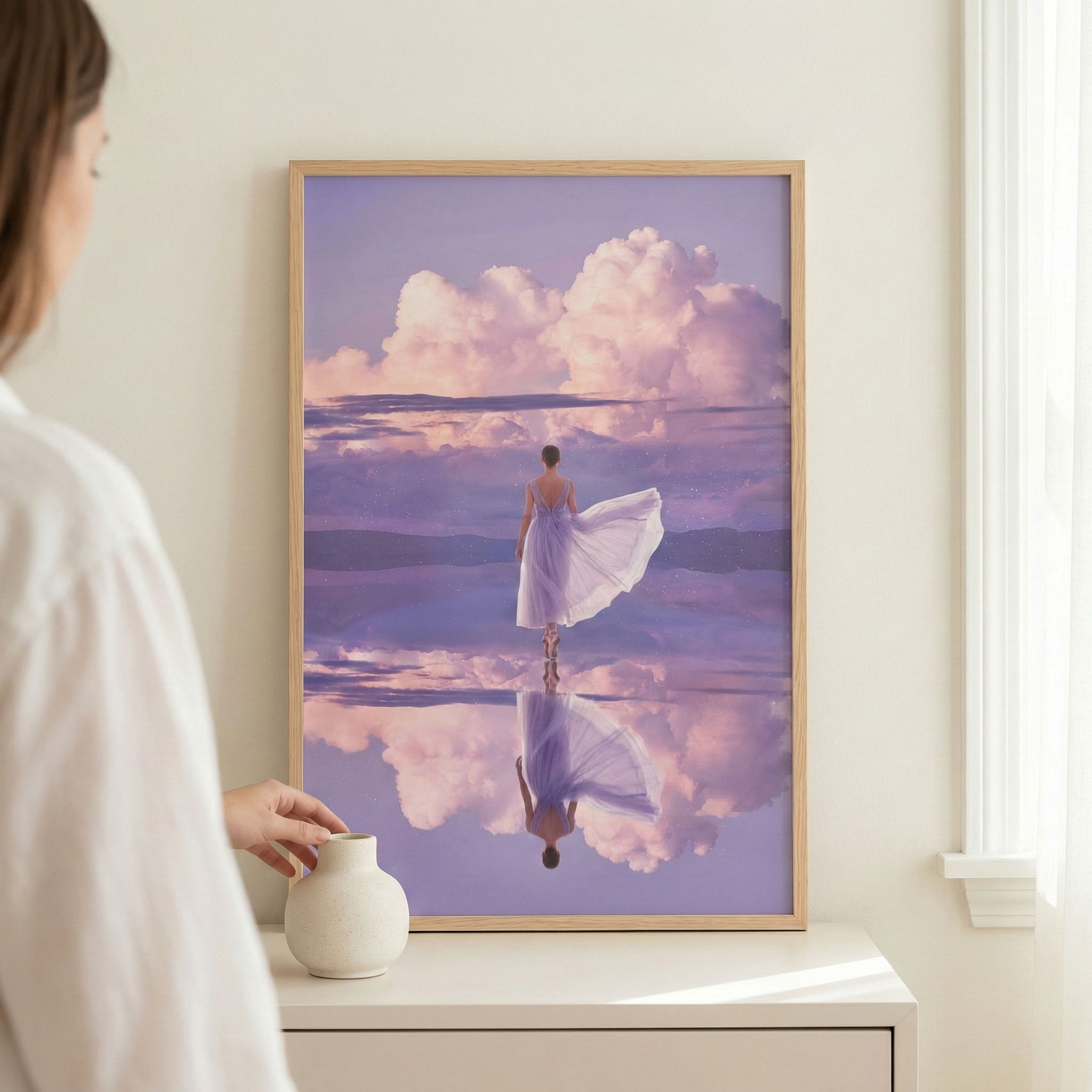 A vertical framed artwork hangs on a neutral wall above a white dresser, next to a small white vase. A person in a white shirt is partially visible on the left. The art depicts an ethereal scene of a lone figure in a flowing lavender dress standing on a perfectly reflective surface. Above, massive, billowy clouds are tinged with hues of pink and purple. Every detail of the person and the dramatic sky is mirrored in the still water below, creating a symmetrical and dreamlike composition.