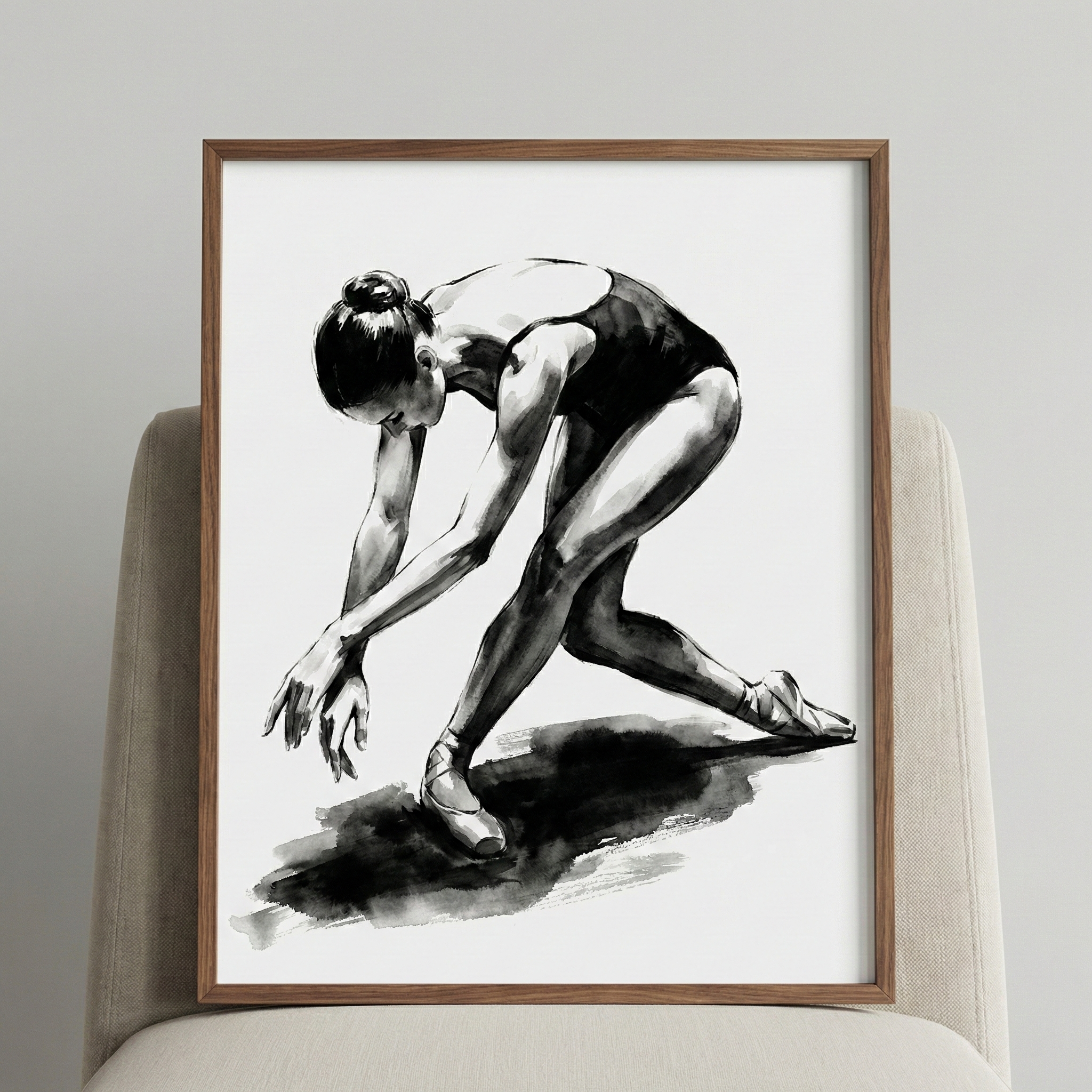 A framed black and white ink-style illustration of a female ballet dancer. The figure is captured in a dynamic, bowed pose, leaning forward with her arms extended toward the ground. She wears a dark leotard and classic ballet shoes. The artwork features bold, expressive strokes and deep shadows, with a prominent dark wash beneath her feet to represent a shadow. The piece is housed in a simple, thin wooden frame and is propped up on a textured, light-colored chair against a neutral wall.