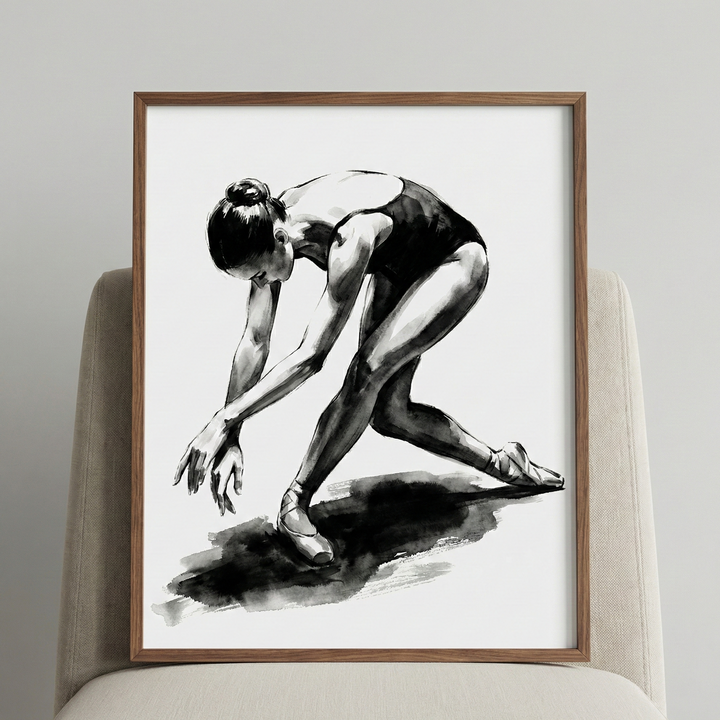 A framed black and white ink-style illustration of a female ballet dancer. The figure is captured in a dynamic, bowed pose, leaning forward with her arms extended toward the ground. She wears a dark leotard and classic ballet shoes. The artwork features bold, expressive strokes and deep shadows, with a prominent dark wash beneath her feet to represent a shadow. The piece is housed in a simple, thin wooden frame and is propped up on a textured, light-colored chair against a neutral wall.