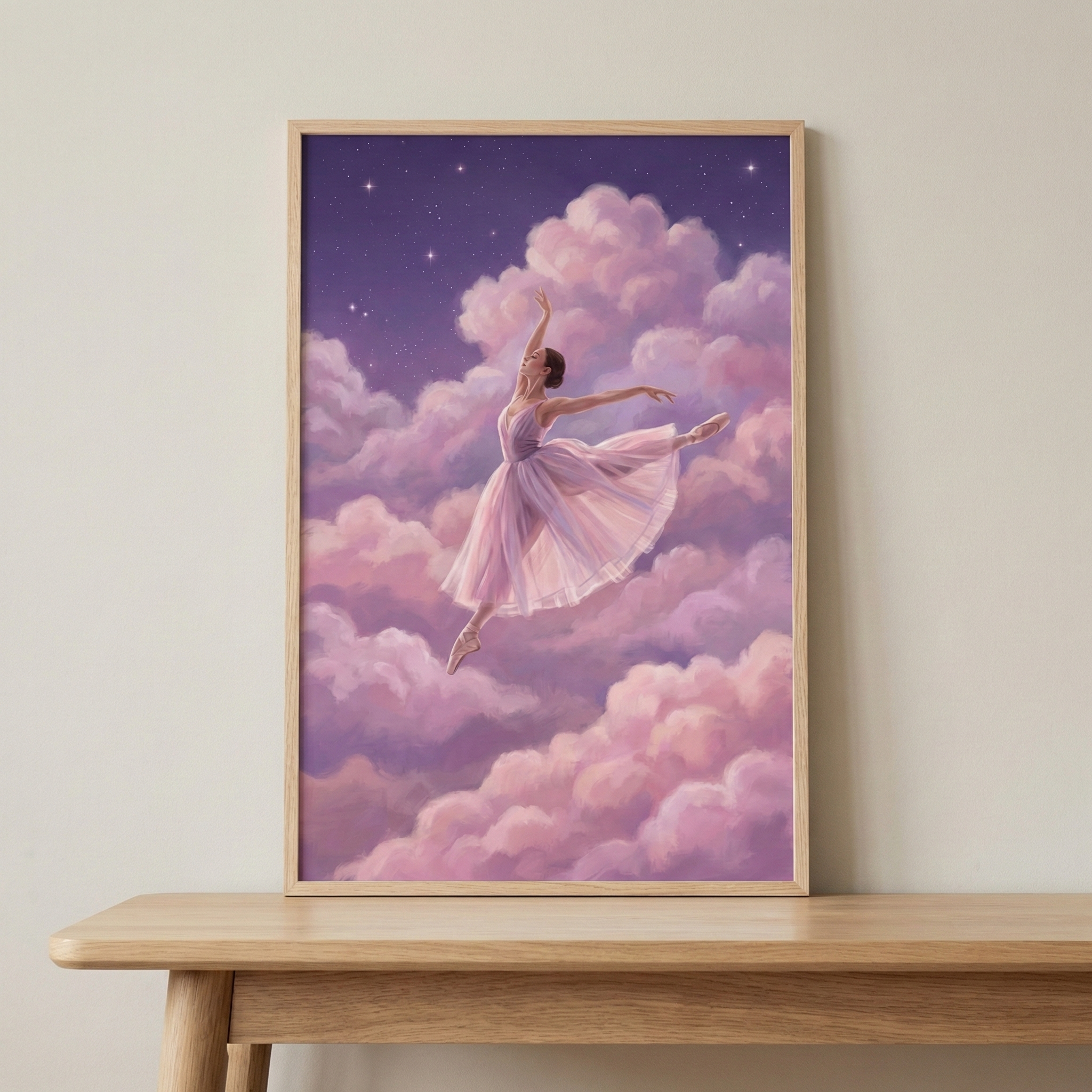 A framed piece of art featuring a ballerina in a graceful leap. She is dressed in a light pink tutu and appears to be dancing among fluffy pink and purple clouds. Above the clouds is a dark purple sky with small, bright stars. The illustration is in a simple light-colored wooden frame, placed on a wooden table against a neutral wall.