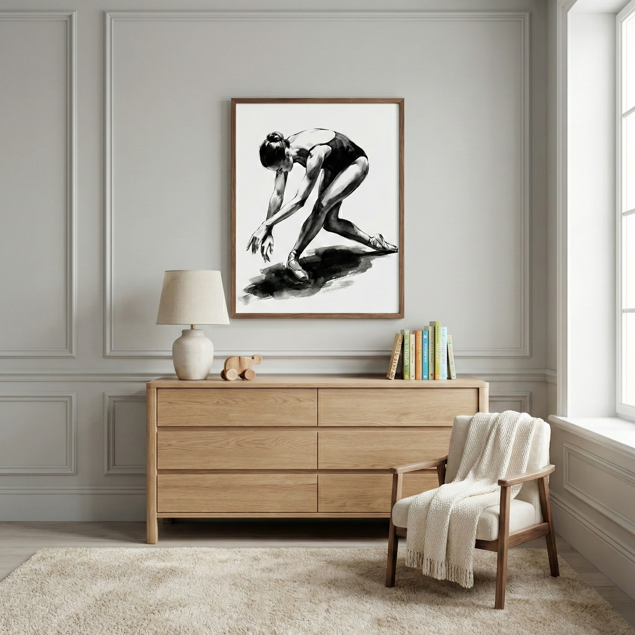 A framed black and white artwork of a ballerina in a graceful pose hangs on a gray paneled wall above a light wood six-drawer dresser. On the dresser sit a neutral lamp, a small wooden animal figure, and a few books. To the right, a wooden armchair with white cushions is draped with a fringed white throw. A plush, light-colored rug covers the floor, and a window on the right provides soft light to the minimalist and serene room.