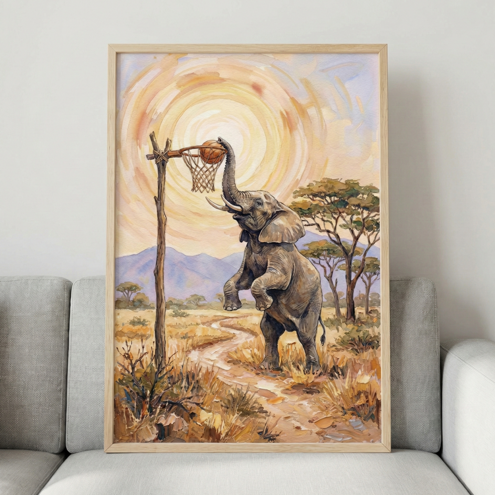 A framed artwork depicts an elephant standing on its hind legs in a savanna landscape. The animal uses its trunk to drop a basketball into a rustic hoop made from a weathered wooden post and a woven net. A winding dirt path leads through dry grass toward distant purple mountains. Large, leafy trees dot the horizon under a sky featuring a massive, swirling golden sun. The piece is encased in a light wood frame and rests against the cushions of a modern gray sofa.
