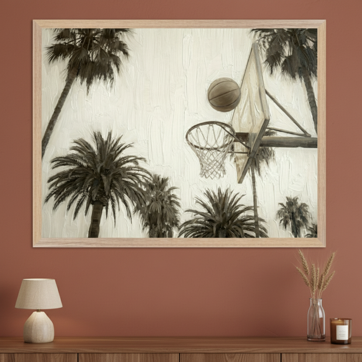 Modern living room with a wooden console table against a burnt orange wall. A large framed, sepia-toned textured print of a basketball hoop, ball, and palm trees hangs above the table. A lamp, vase with wheat, and candle sit on the table.