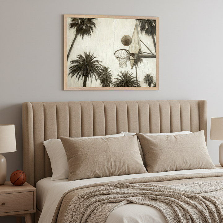 Modern bedroom with a beige upholstered bed and matching pillows. A large framed print of a black and white basketball hoop and palm trees hangs centered above the bed. A small wooden nightstand with a lamp and a basketball is to the left.
