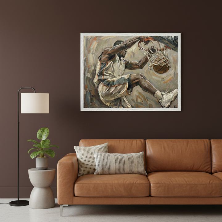 Modern living room with a tan leather sofa against a dark brown wall. A white-framed painting of a basketball player dunking in a brushstroke style hangs centered above the sofa. A floor lamp and plant on a side table are to the left.