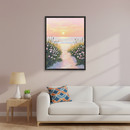 Living room with a white sofa and a wooden side table. A framed painting of a beach path leading to the ocean at sunset, flanked by grass and pink and white flowers, hangs centered above the sofa.