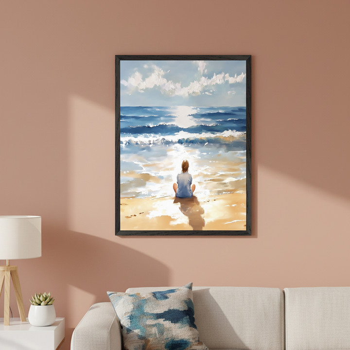 Modern living room with a light-colored sofa and side table. A large black-framed painting hangs centered above the sofa, depicting a person sitting on a sunlit beach facing the ocean under a blue, cloudy sky in an impressionistic style.