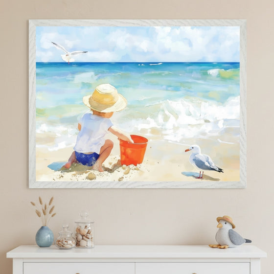 A child kneels on the beach facing the ocean, wearing a straw hat and playing with a red bucket in the sand, while a seagull watches close by. The framed painting hangs above a dresser with soft coastal decorations, evoking a peaceful seaside scene.