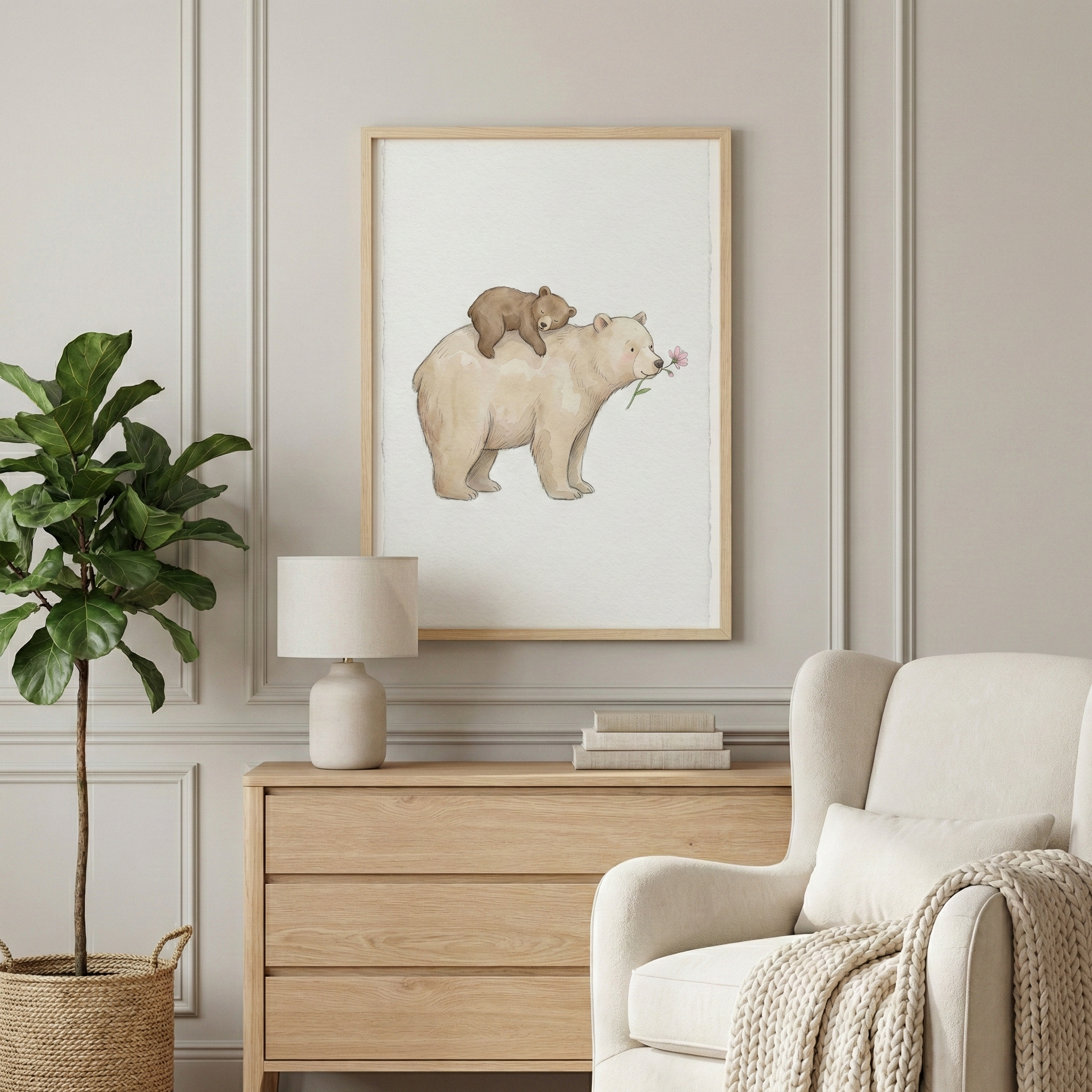 A bright, neutral room interior features a framed art piece on a wall with decorative molding. The art illustrates a large, cream-colored bear with a small brown cub resting on its back; the larger animal holds a soft pink flower in its mouth. Below the frame sits a light wood dresser holding a simple white lamp and a few books. A tall green plant in a woven basket stands to the left, while an off-white armchair with a cozy throw blanket and pillow is positioned on the right.
