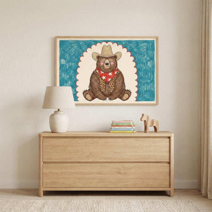 Charming cowboy bear wall art hung above a light wood dresser with a neutral lamp and stack of children’s books, creating a soft, cozy atmosphere. The framed artwork brings a warm and whimsical focal point to nursery or kids’ room wall art.