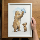 A white-framed watercolor illustration rests on a light wood surface, held by a hand. The artwork depicts a large brown bear and a small cub standing on their hind legs against a minimalist white background with a soft splash of blue. The larger bear holds a light blue book high above its head. The cub stands below, reaching its paws upward toward its parent and the book with an eager expression. The piece features soft color washes and delicate ink outlines.