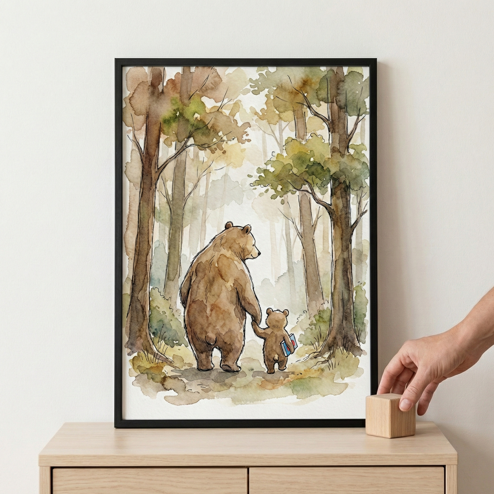 A framed watercolor illustration depicts a large brown bear and a small bear cub walking hand-in-hand away into a forest. The cub carries a blue book under its arm. They are surrounded by tall trees with soft green and brown leaves, rendered in a gentle, atmospheric style. The art piece is set in a black frame and sits on a wooden sideboard, with a person's hand visible to the side placing a small wooden block.