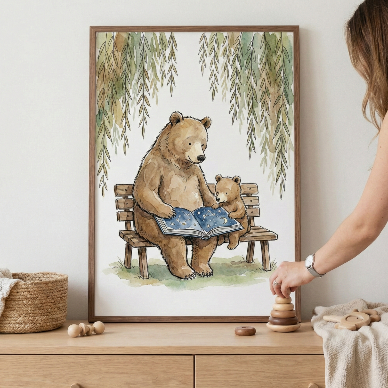 An illustration in a wooden frame shows a large brown bear and a small cub sitting together on a wooden bench. The adult bear holds an open book featuring a dark blue sky with white stars. Wispy green branches hang over the bears from above. The framed artwork is placed on a light wood dresser next to a woven basket and small wooden toys. On the right, a person's hand is seen stacking wooden rings on the dresser's surface.