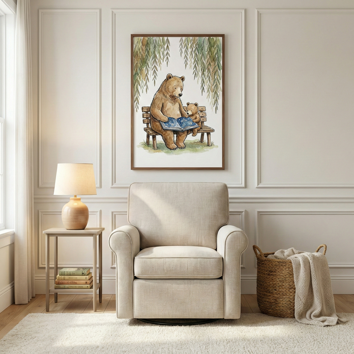 A cozy reading corner features a vertical watercolor illustration of a large brown bear and a small bear cub sitting together on a wooden bench, looking at an open book. Weeping willow branches hang over them. The artwork is in a thin brown frame and hangs on a light-colored wall with decorative rectangular molding. Below the art is a beige upholstered swivel glider chair with rounded arms. To the left, a light wood side table holds a ceramic lamp with a glowing shade and a small stack of books. 