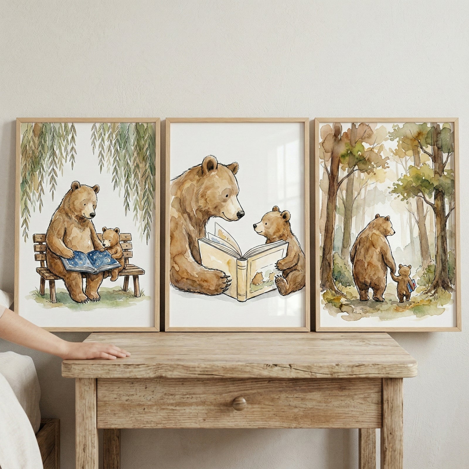 A series of three light-wood framed watercolor illustrations displayed on a light-colored wall above a rustic wooden side table. The whimsical artwork features an adult bear and its cub in various endearing scenes. On the left, they sit on a park bench under willow trees reading a book with a starry cover. The center piece shows a close-up of them reading together. On the right, they walk through a forest hand-in-hand. A person's arm rests on the table's edge in the foreground.