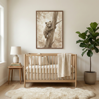 A neutral-toned nursery features a light wood crib centered against an off-white wall. Above the crib hangs a dark wood frame containing an illustration of a small bear cub climbing a tree in sepia-toned watercolor style. A textured knit blanket is draped over the crib's side. To the left is a wooden side table with a cream lamp and a stack of books. To the right is a large potted leafy green plant. A soft, white sheepskin rug lies on the light wood floor in the foreground.
