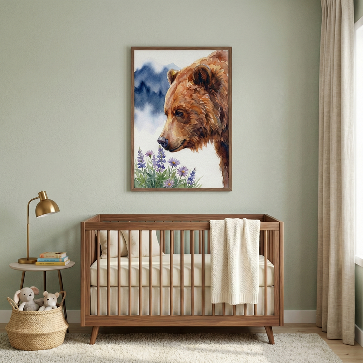 A serene nursery features a mid-century modern wooden crib centered against a soft sage green wall. Above the crib hangs a large framed watercolor-style illustration depicting a brown bear gazing at purple wildflowers with distant blue mountains. A cream-colored blanket is draped over the crib's edge. To the left, a gold desk lamp sits on a small round table next to a stack of books. A woven basket with two stuffed animals rests on the plush white rug below.