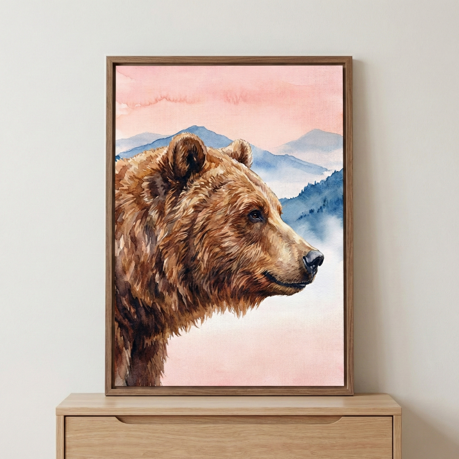 A watercolor illustration of a grizzly bear's head in profile, highlighting the detailed texture of its thick brown fur. The bear is set against a serene background of misty blue mountain ranges and a soft pink and peach-colored sky. The artwork is created with delicate watercolor washes and soft brushwork, giving it a peaceful, nature-inspired feel. The entire scene is shown inside a simple, light wood frame, positioned on top of a light wooden cabinet against a clean, neutral-toned wall.