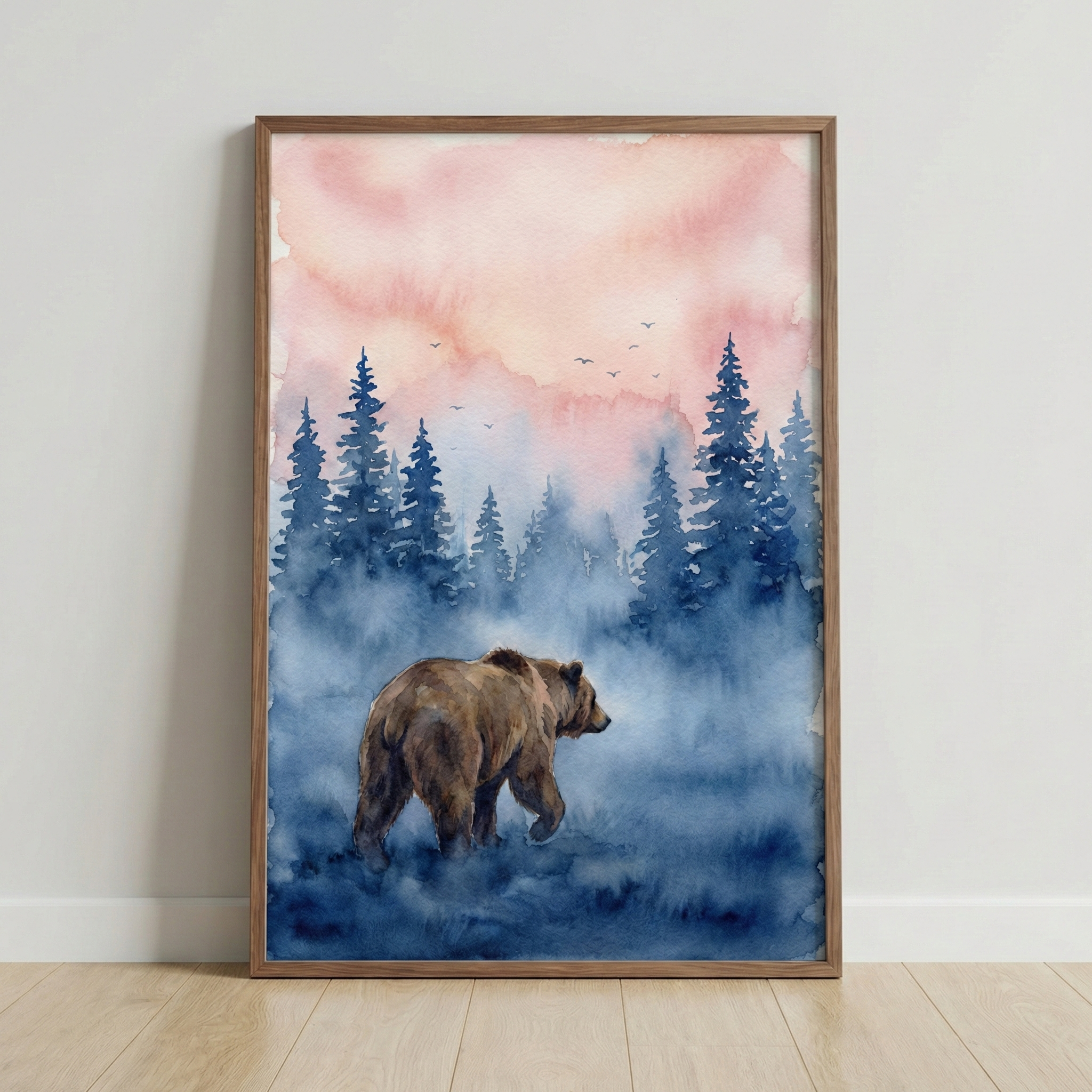 A framed watercolor artwork shows a brown bear walking through blue mist in a forest of tall pine trees. The sky above fades from soft peach to pale pink with small birds flying in the distance, creating a calm woodland scene. The piece leans against a light wall on a wooden floor, highlighting its cozy nature inspired style.