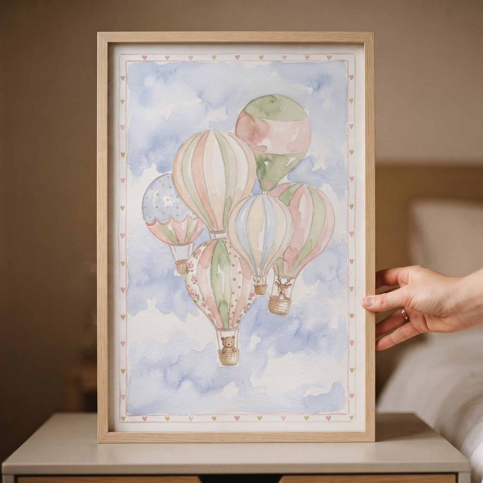 Hand holding framed bear hot air balloon wall art with watercolor pastel balloons and a teddy bear in a blue cloudy sky.