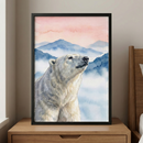 A serene watercolor-style illustration of a polar bear is displayed in a slim black frame. The bear is shown from the shoulders up, looking upward and to the side with a calm expression. Its fur is captured with soft washes of white, cream, and grey. Behind the bear, a landscape of rolling blue mountains rises into a soft pink and peach-colored sky, with white mist settled in the valleys. This framed artwork sits on a light-colored wooden nightstand next to a corner of a bed, creating a peaceful atmosphere.