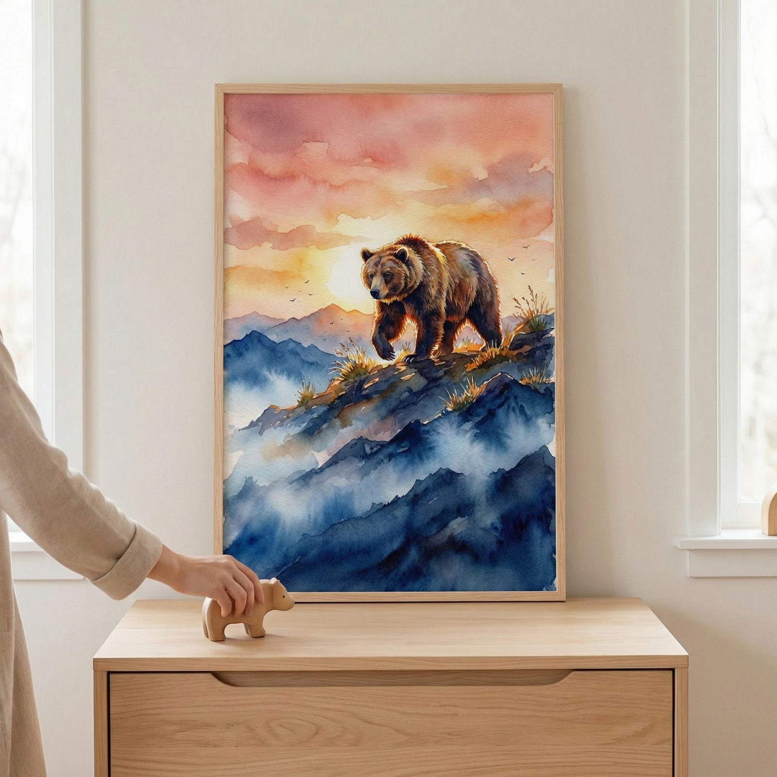 A vertical watercolor-style art piece in a light wood frame depicts a large brown bear walking along a rocky mountain ridge. The background features a sunset with clouds in shades of pink, orange, and yellow. Below the bear, rugged mountains are covered in deep blue tones and white mist. The frame is resting on a light wooden dresser next to a window. In the foreground, a person's hand reaches for a small wooden bear figurine on the dresser's surface. The room has a soft and peaceful atmosphere.