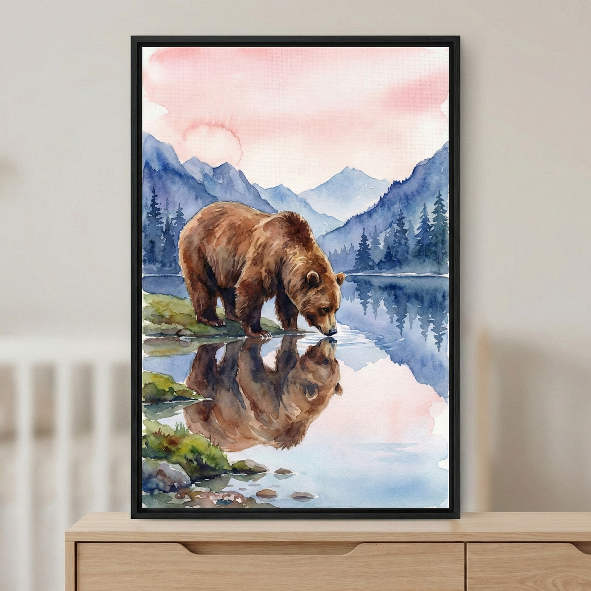A serene watercolor artwork depicting a large brown bear at a lake's edge. The bear is captured in a profile view, leaning down to drink from the clear water, which perfectly mirrors its form. The landscape features rolling blue mountain peaks and a dense forest of evergreen trees under a soft, pastel pink and orange sky. This piece is presented in a slim black frame, resting on a light wood dresser. The style is defined by its fluid brushwork and gentle color palette.