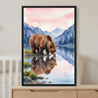 A serene watercolor artwork depicting a large brown bear at a lake's edge. The bear is captured in a profile view, leaning down to drink from the clear water, which perfectly mirrors its form. The landscape features rolling blue mountain peaks and a dense forest of evergreen trees under a soft, pastel pink and orange sky. This piece is presented in a slim black frame, resting on a light wood dresser. The style is defined by its fluid brushwork and gentle color palette.