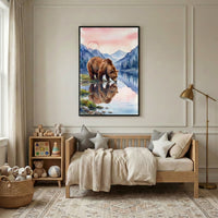 Framed watercolor painting of a brown bear standing at the edge of a calm mountain lake with its reflection visible in the water, hung above a wooden daybed in a cozy neutral toned nursery. The room features soft beige bedding, star shaped pillows, a knit throw blanket, a brass floor lamp, and a small shelf with stuffed animals and wooden toys, creating a warm and inviting space.